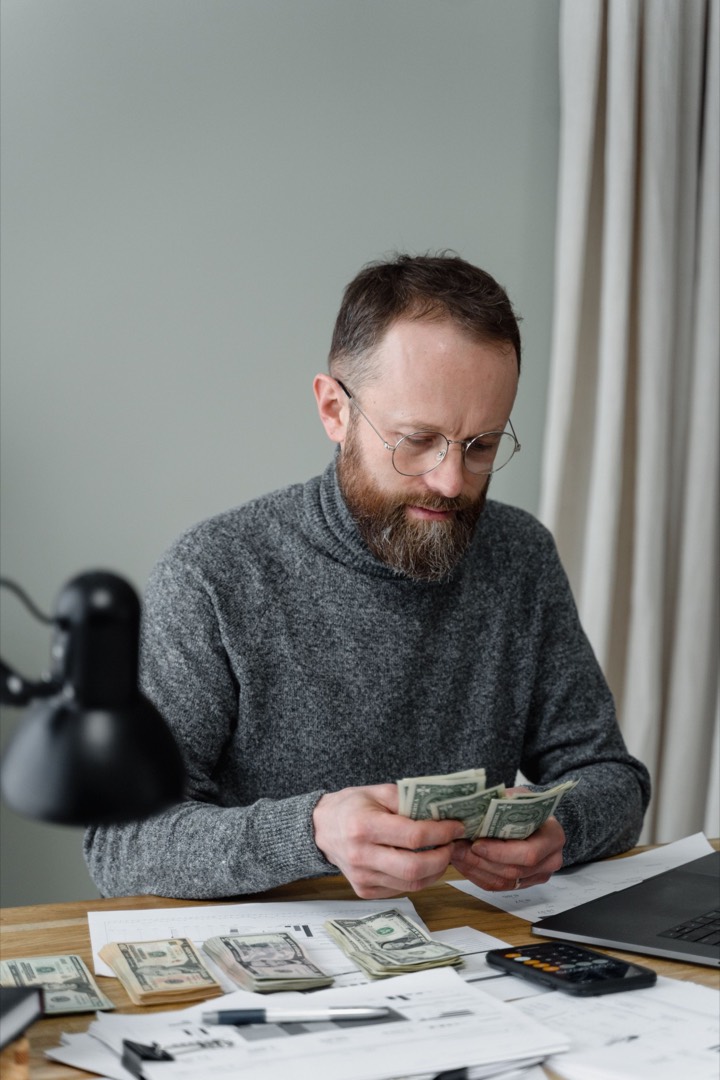 A man in glasses and a gray sweater counting cash at a desk covered with papers, a calculator and a laptop